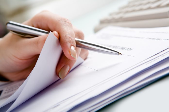 Close-up of hand holding pen with paper over pile of documents