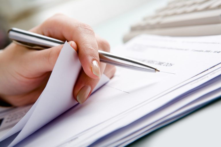 Close-up of hand holding pen with paper over pile of documents