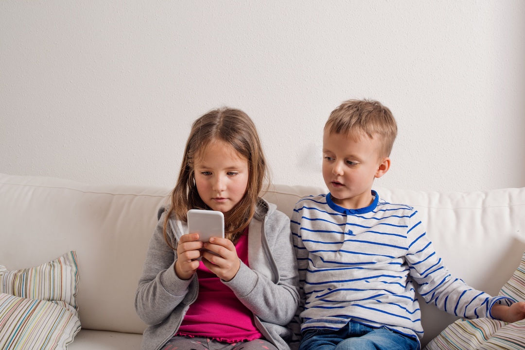 Little girl and boy sitting on sofa with a smart phone at home. Happy children playing indoors, copy space